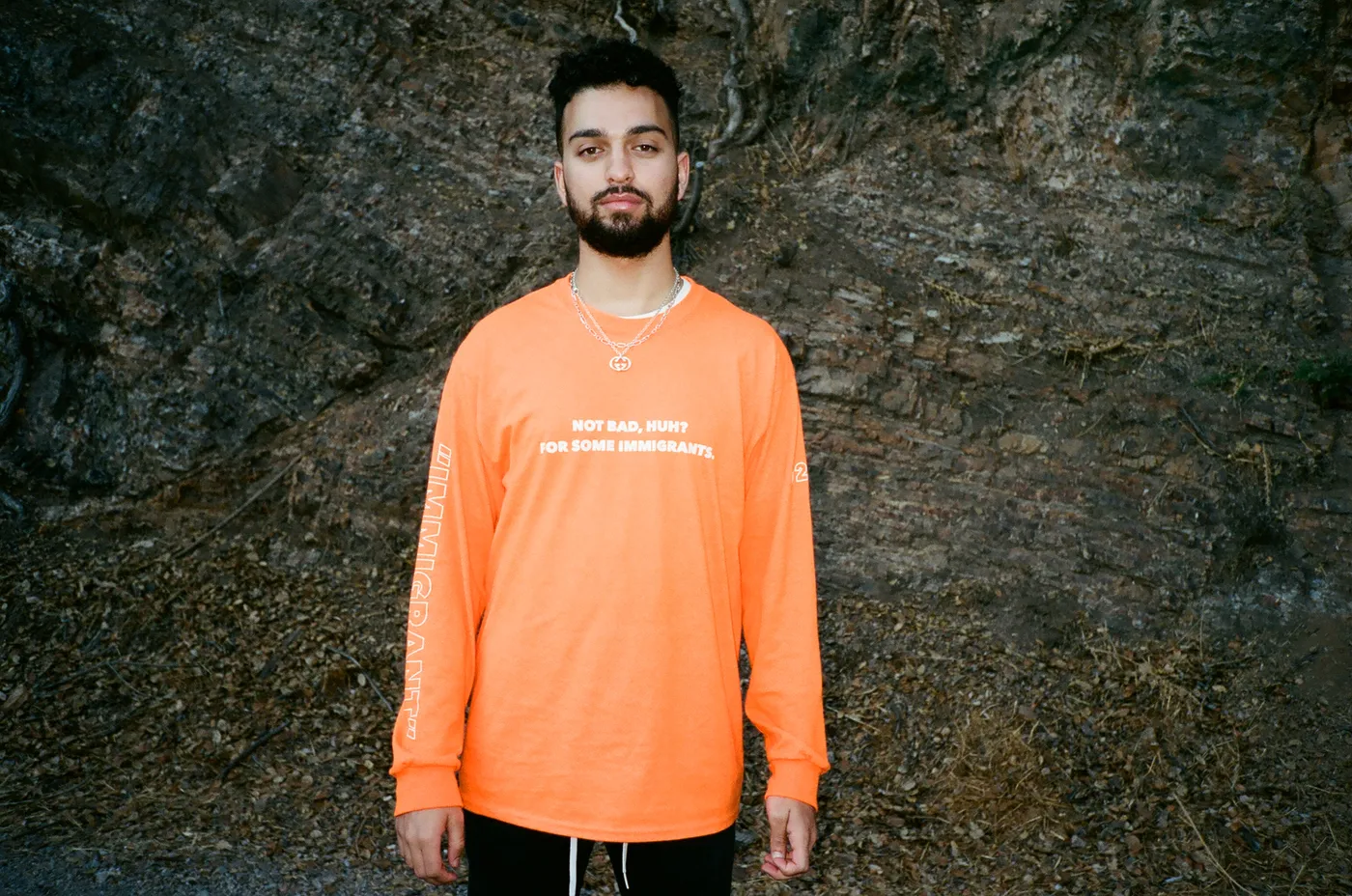 Model standing against a rock face in the Not Bad, Huh? shirt, chest print reading NOT BAD, HUH? FOR SOME IMMIGRANTS